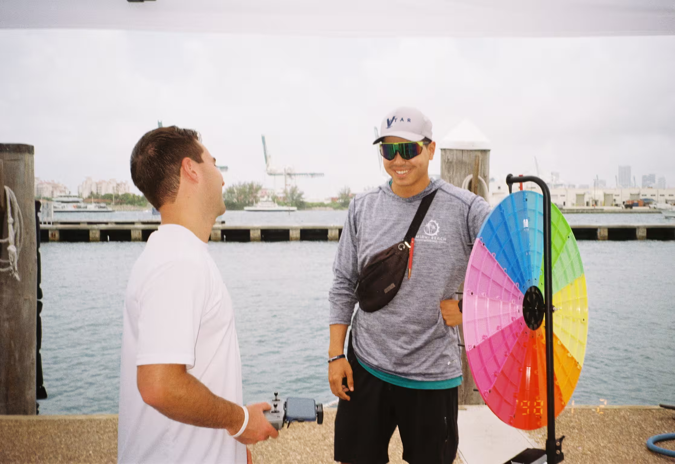 Two men talking on a marina dock, one holding a drone controller, a prize wheel and the waterfront behind them.
