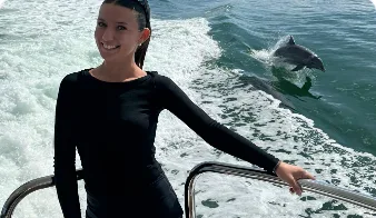 Anna Dyniszkiewicz at a yacht's stern rail, a dolphin breaking the wake behind her.
