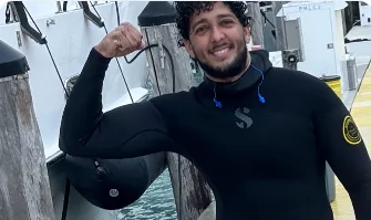 Orlando Noriega in a wetsuit on a dock, a dive hood over his arm.